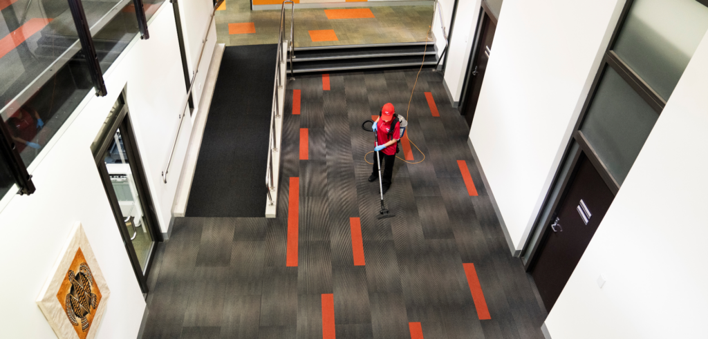 A commercial cleaner vacuuming the carpeted hallway of a large enterprise building from an overhead perspective.